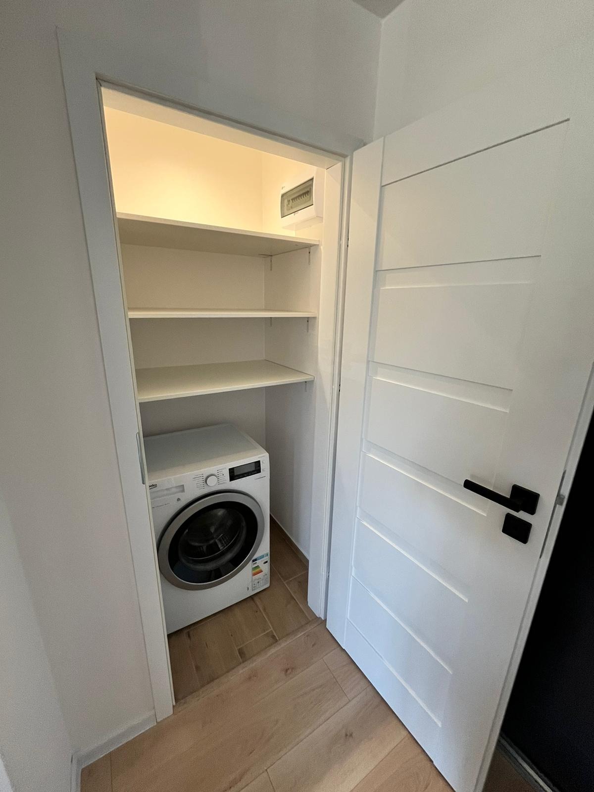 Biała pralka w zabudowie z półkami, widoczna przez otwarte drzwi z czarną klamką. Jasne wnętrze, drewnopodobna podłoga. Minimalistyczny design.