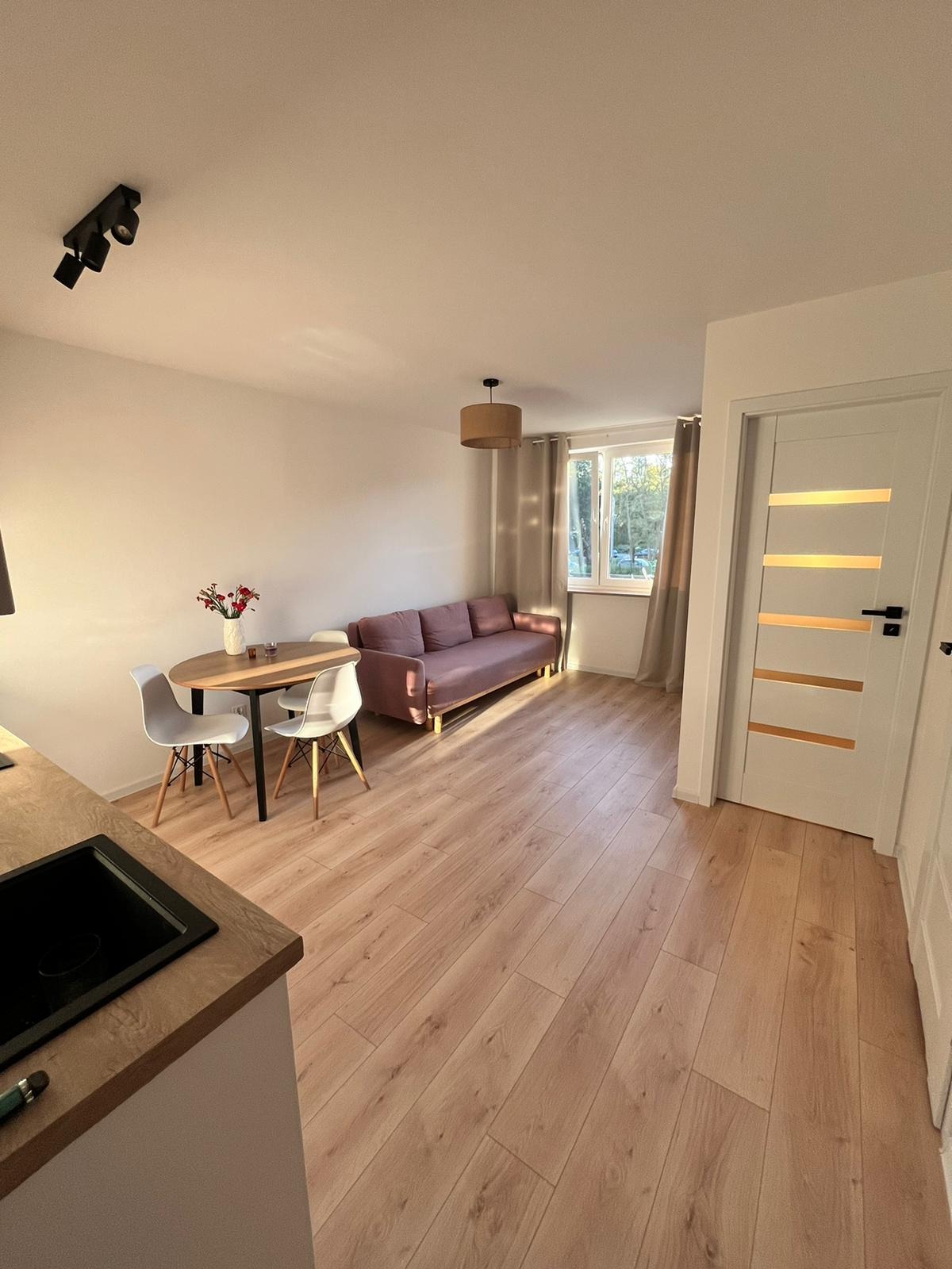 Jasne panele podłogowe w pokoju z sofą, stołem i białymi krzesłami. Widoczne drzwi z poziomymi przeszkleniami i fragment aneksu kuchennego.