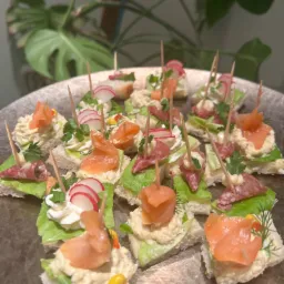 Zbliżenie na srebrnej tacy z r&oacute;żnorodnymi, apetycznymi kanapeczkami na bazie pieczywa, sałaty, łososia, rzodkiewki i salami, spiętymi wykałaczkami, z rośliną w tle.