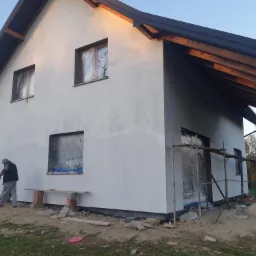 Dwukondygnacyjny dom w trakcie nakładania pierwszej warstwy szarego tynku, okna zabezpieczone folią, widoczne rusztowanie i pracownik w szarym kombinezonie.