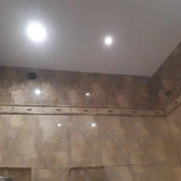 Beżowe płytki imitujące marmur z dekoracyjnym paskiem w górnej części ściany łazienki, widoczne oświetlenie sufitowe punktowe i fragment wentylacji.