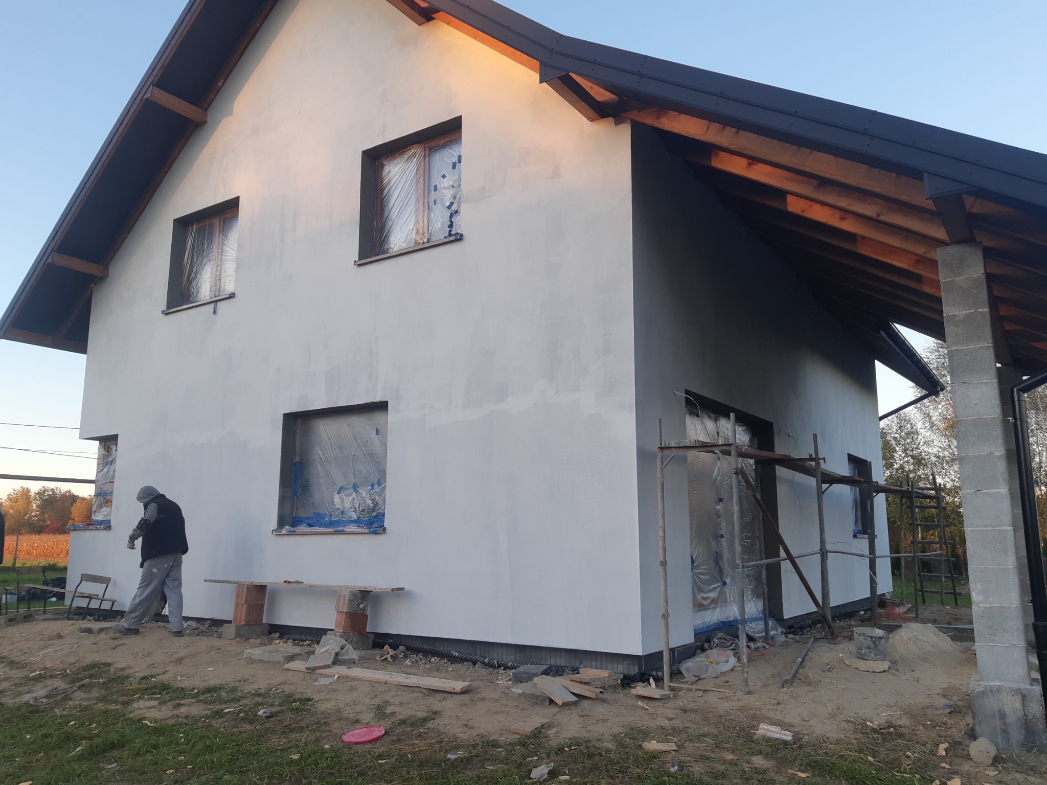 Dwukondygnacyjny dom w trakcie nakładania pierwszej warstwy szarego tynku, okna zabezpieczone folią, widoczne rusztowanie i pracownik w szarym kombinezonie.