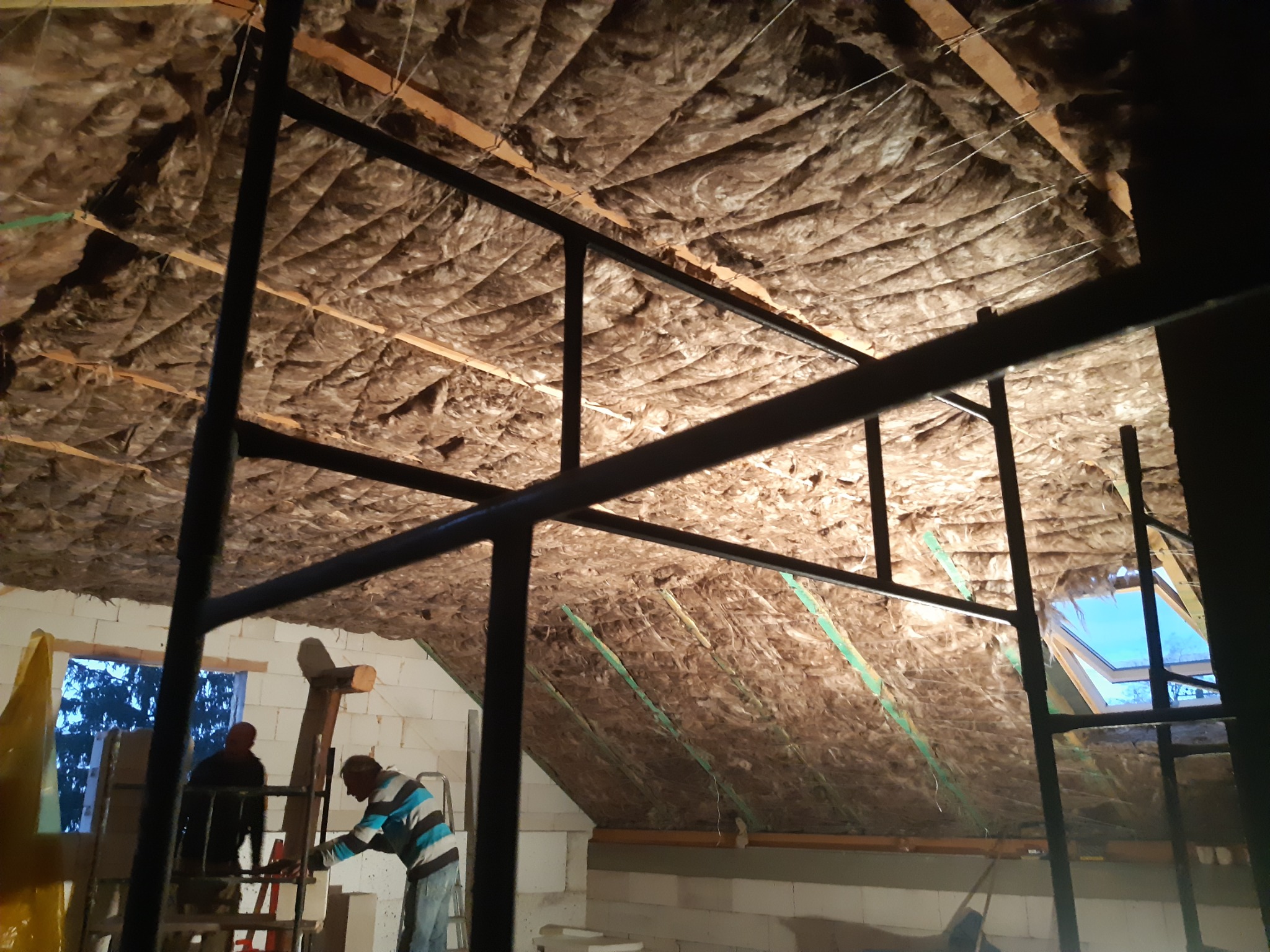 Ocieplenie poddasza wełną mineralną widoczne przez stalową konstrukcję rusztowania, dwóch pracowników na drabinie w tle, okno dachowe w oddali.