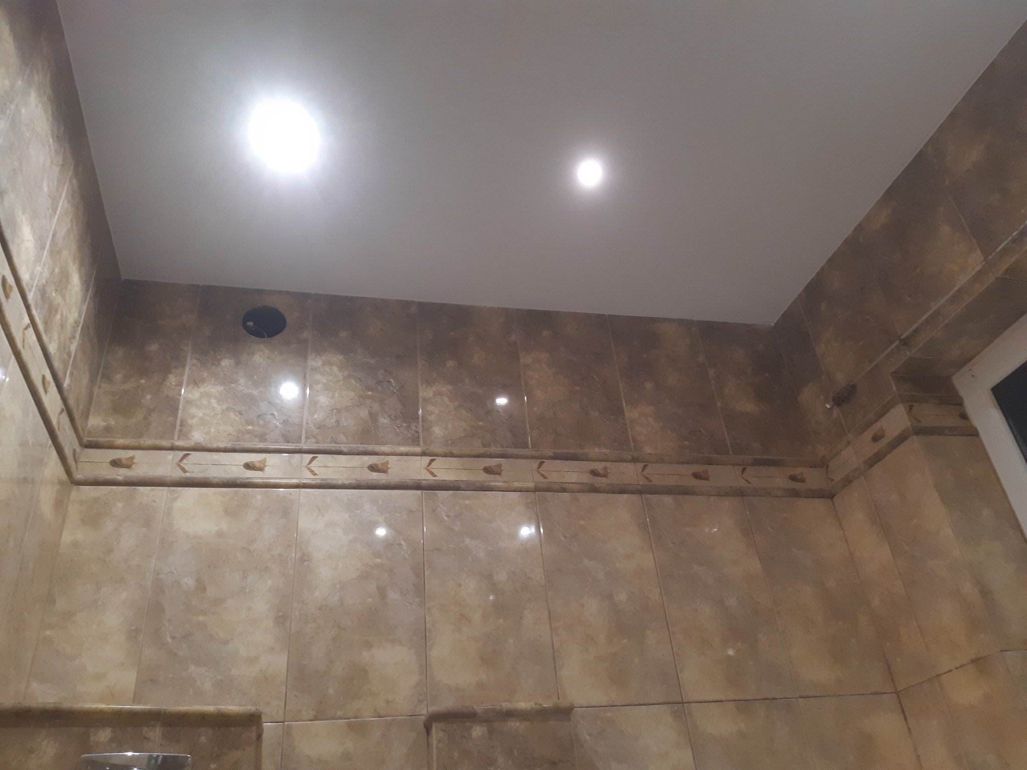 Beżowe płytki imitujące marmur z dekoracyjnym paskiem w górnej części ściany łazienki, widoczne oświetlenie sufitowe punktowe i fragment wentylacji.