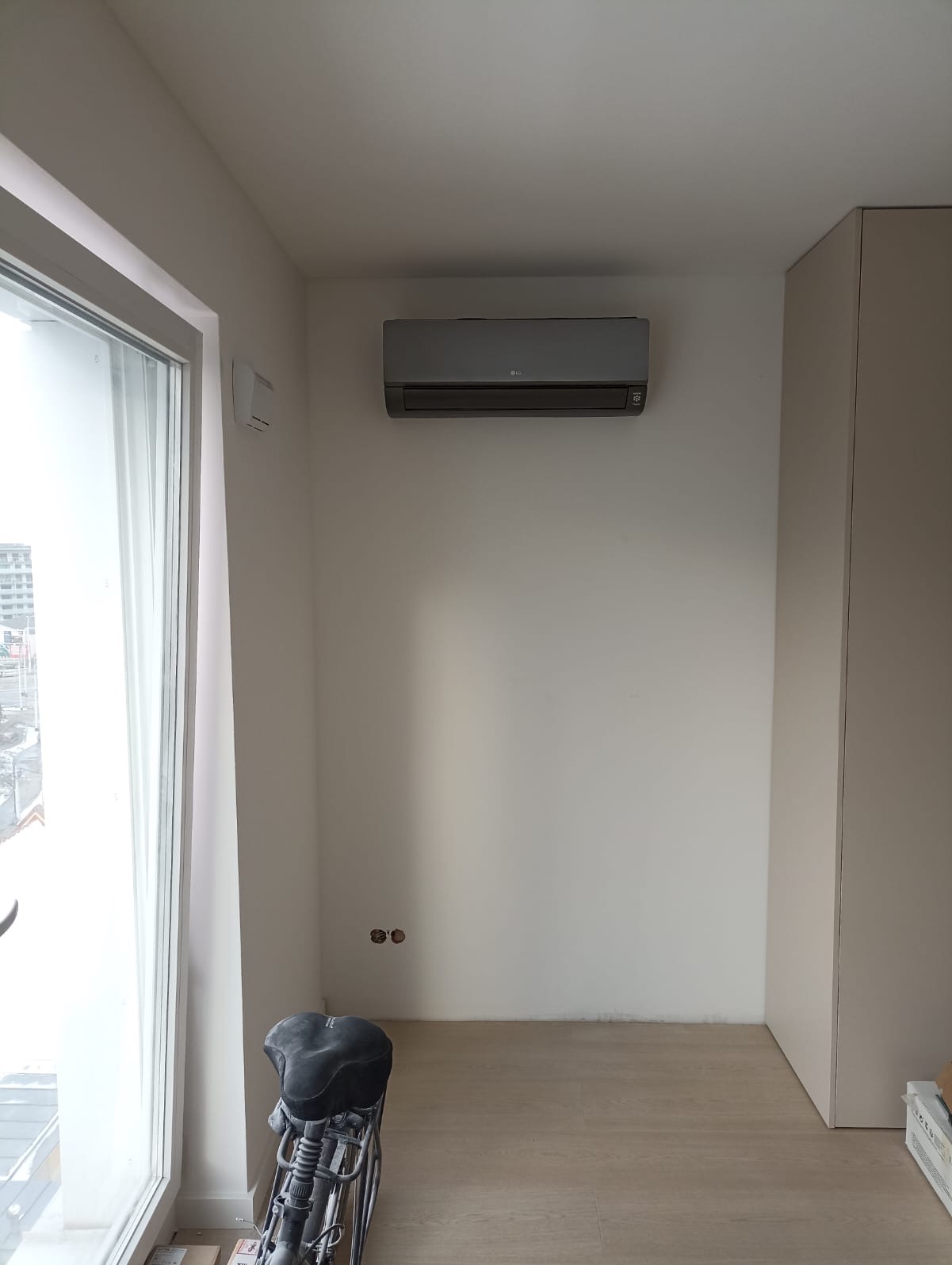 Szara jednostka klimatyzacji LG Artcool Black Mirror zamontowana na białej ścianie w jasnym pomieszczeniu z dużym oknem i fragmentem roweru na podłodze.