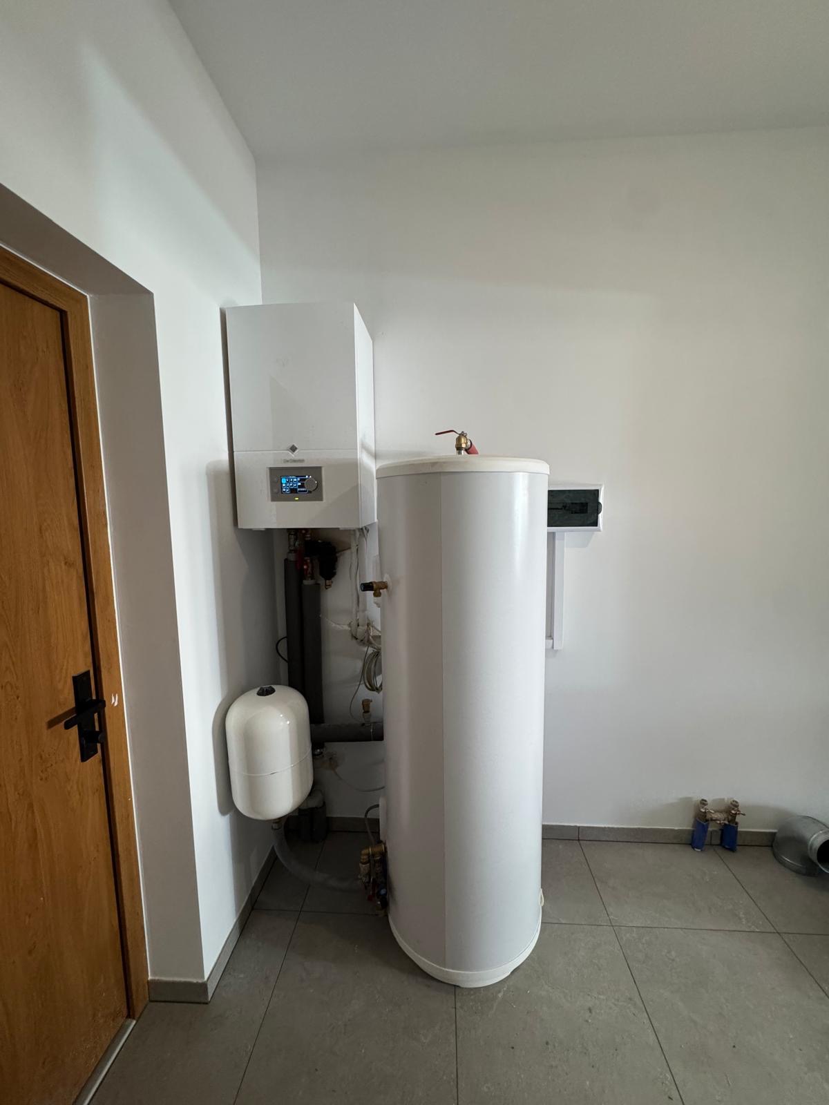 Biała pompa ciepła De Dietrich z zbiornikiem w jasnym pomieszczeniu, widoczne rury i elementy instalacji. Ujęcie z niskiego kąta, drzwi po lewej stronie kadru.