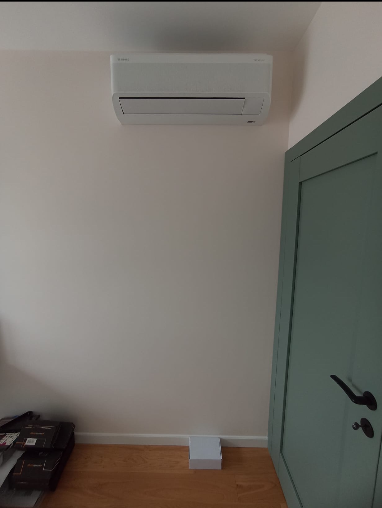 Biała jednostka wewnętrzna klimatyzacji Samsung Wind-Free Avant zamontowana na ścianie w jasnym pomieszczeniu, obok zielonych drzwi. Widoczny fragment podłogi.