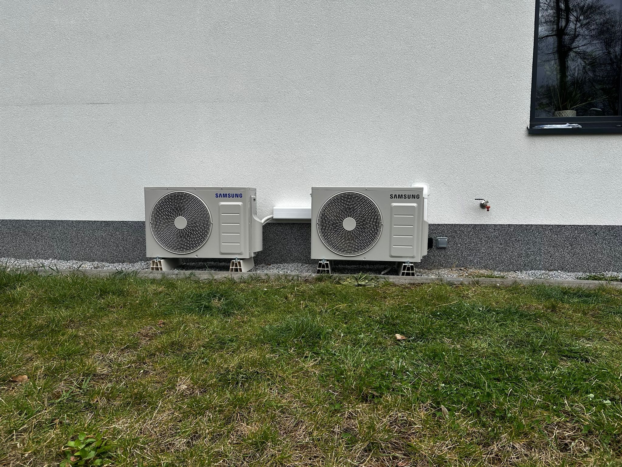 Dwa zewnętrzne agregaty klimatyzacji Samsung Wind-Free Avant, zamontowane na niskich podstawach przy ścianie budynku z oknem i trawnikiem. Widok z poziomu gruntu.