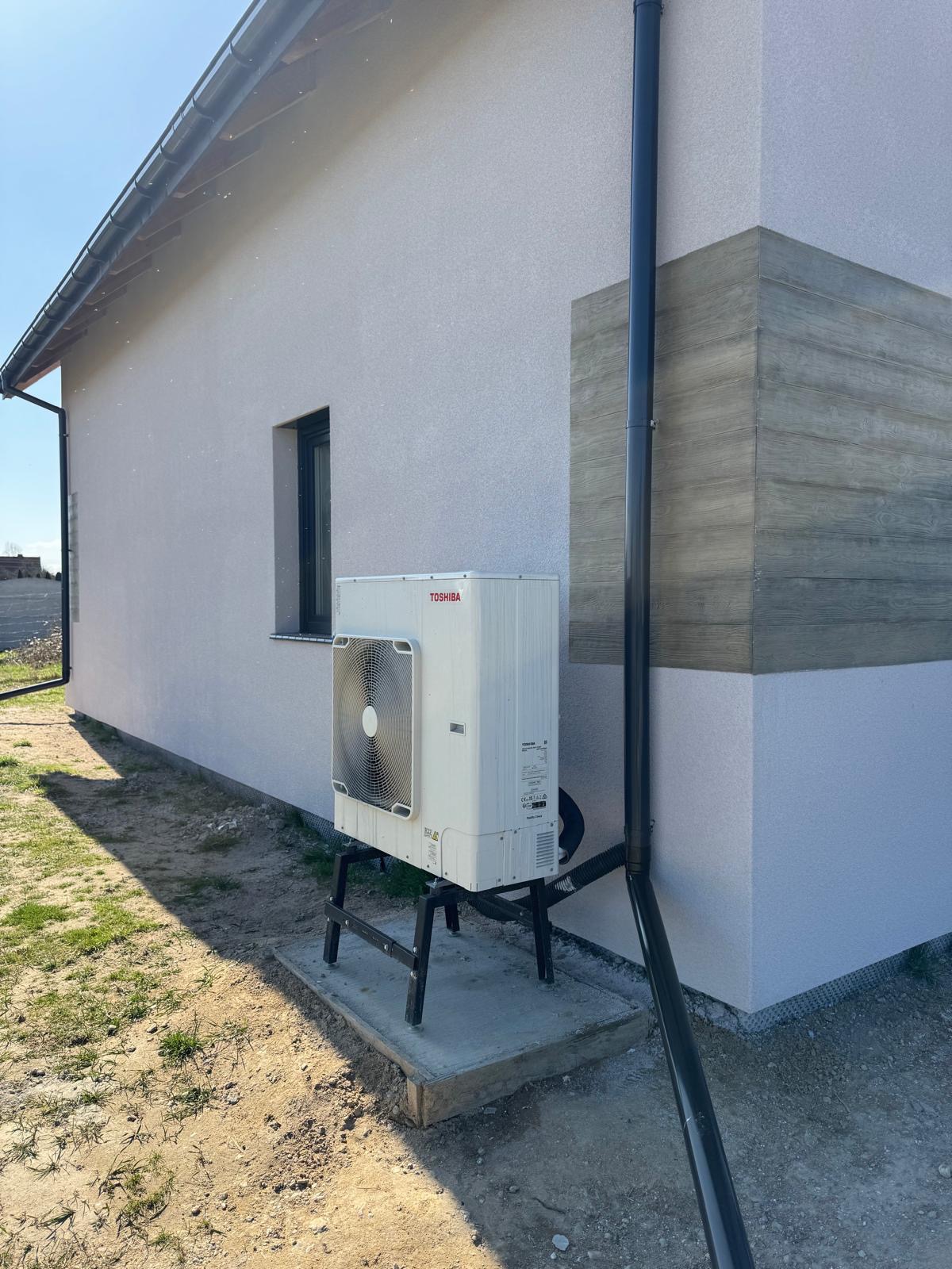 Biała pompa ciepła Toshiba zamontowana na zewnątrz domu jednorodzinnego, widoczna rynna spustowa i fragment elewacji z oknem. Ujęcie z dołu, w słoneczny dzień.