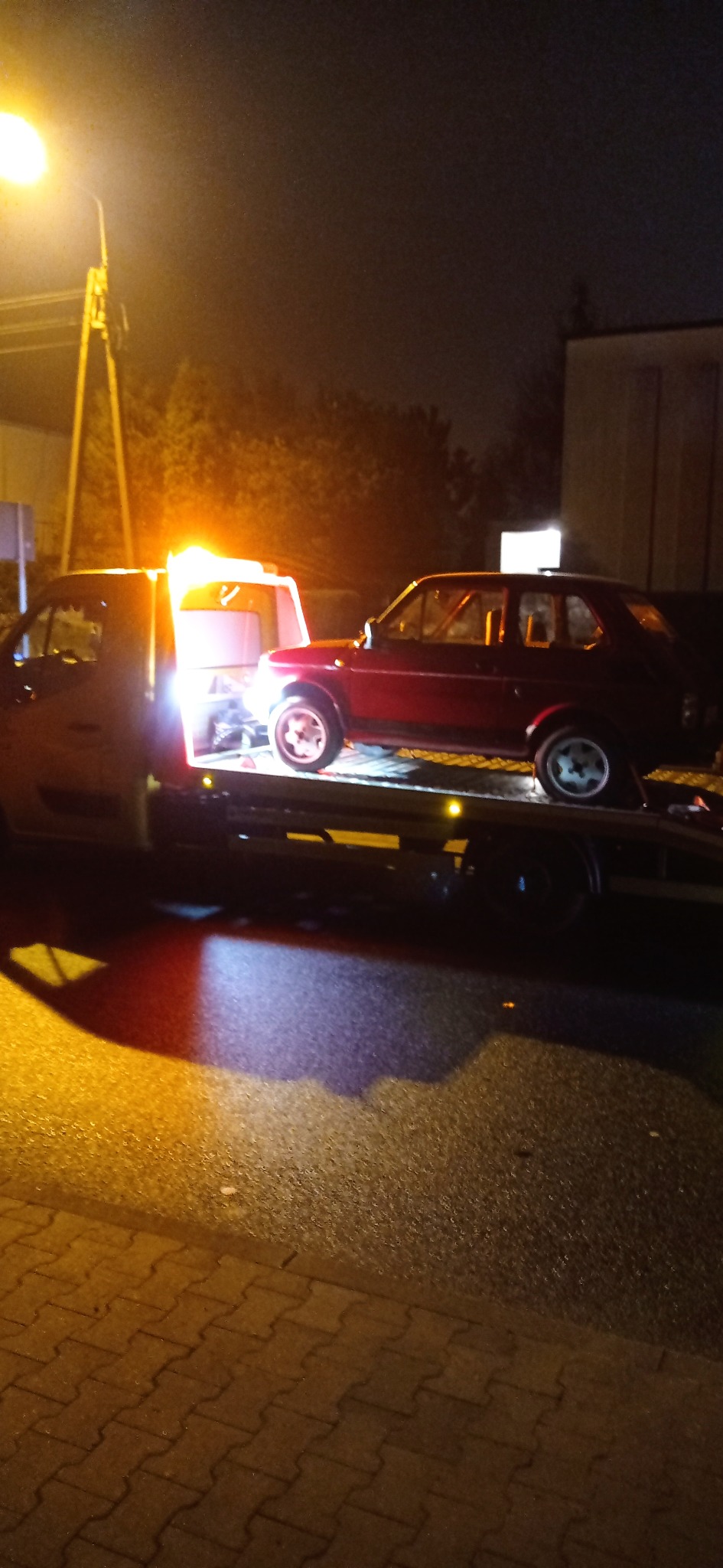 Czerwony samochód osobowy, prawdopodobnie Fiat, transportowany na lawecie w nocy, oświetlony przez światła lawety i uliczną lampę.