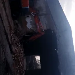 Pomarańczowa koparka wyburza ceglany budynek, widoczne gruzy i fragmenty konstrukcji, ciemne tło.