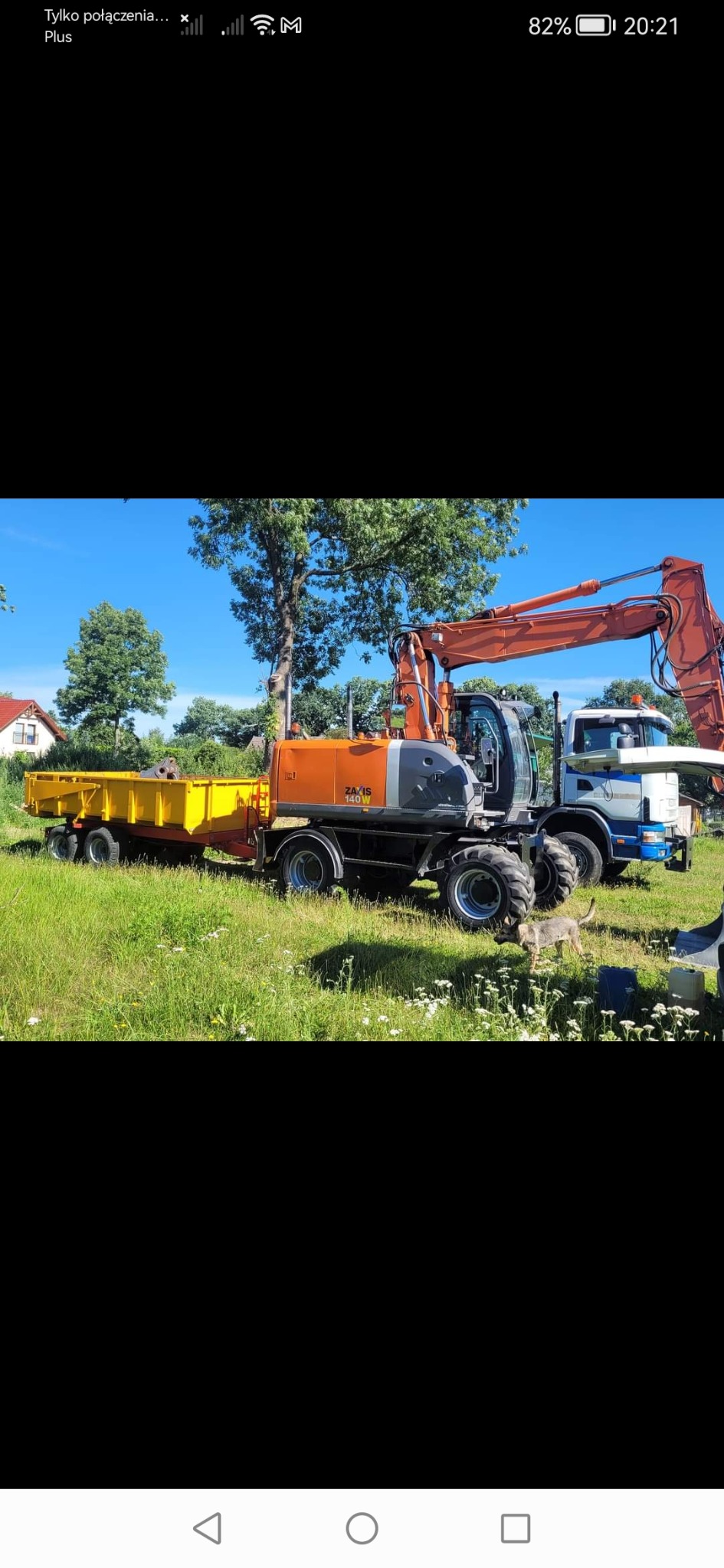 Pomarańczowa koparka kołowa ZA/IS 140W z żółtą przyczepą na trawiastym terenie, obok zaparkowany biało-niebieski samochód ciężarowy, w tle drzewa i dom.