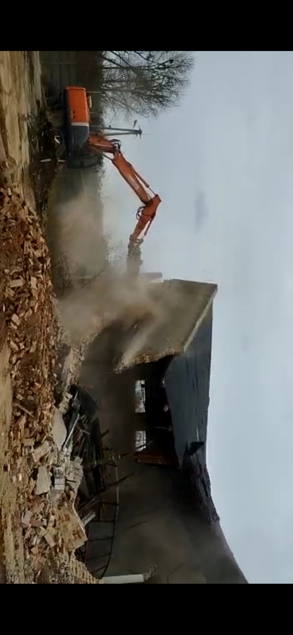 Pomarańczowa koparka z długim ramieniem kruszy resztki budynku, unoszący się pył, gruzowisko cegieł i elementów konstrukcyjnych na pierwszym planie, pochmurne niebo w tle.