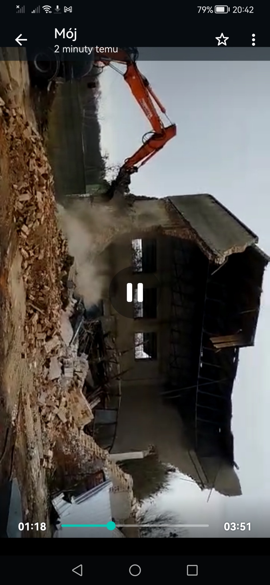 Pomarańczowa koparka z wysięgnikiem kruszy pozostałości budynku podczas rozbiórki, unosząc tumany kurzu.