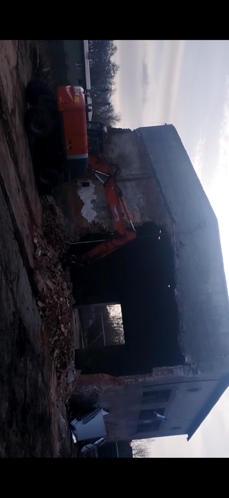 Pomarańczowa koparka wyburza ceglany budynek, widoczne gruzy i fragmenty konstrukcji, ciemne tło.