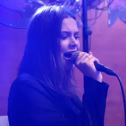 Wokalistka z mikrofonem śpiewa na scenie w niebieskim oświetleniu, widoczny kabel mikrofonowy.