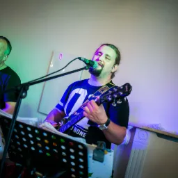 Dwaj mężczyźni grający muzykę na żywo: jeden na keyboardzie, drugi na gitarze, obaj śpiewają do mikrofonów, w tle dekoracyjne elementy architektoniczne.