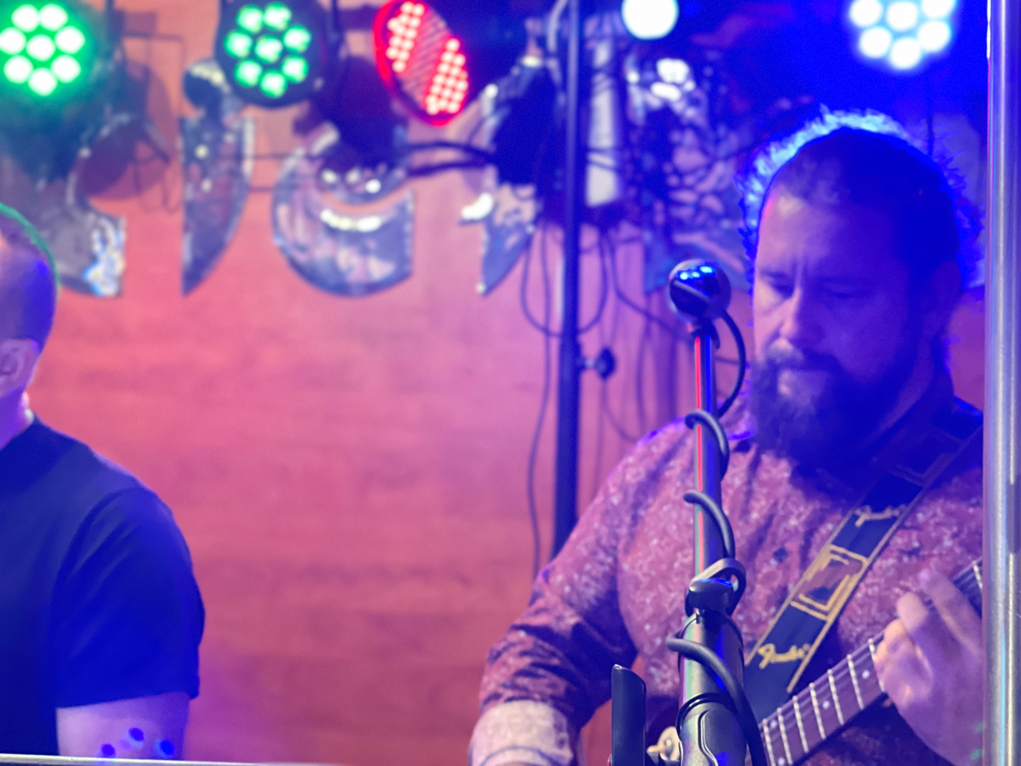 Gitarzysta z brodą gra na elektrycznej gitarze Fender na scenie z oświetleniem scenicznym, widoczny mikrofon i drugi muzyk z lewej strony kadru.