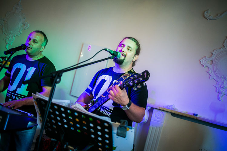 Dwaj mężczyźni grający muzykę na żywo: jeden na keyboardzie, drugi na gitarze, obaj śpiewają do mikrofonów, w tle dekoracyjne elementy architektoniczne.