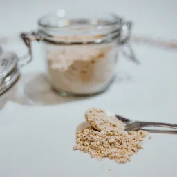 Jasny proszek w szklanym słoiku z metalowym zamknięciem, obok łyżeczka z wysypanym proszkiem na białym tle.