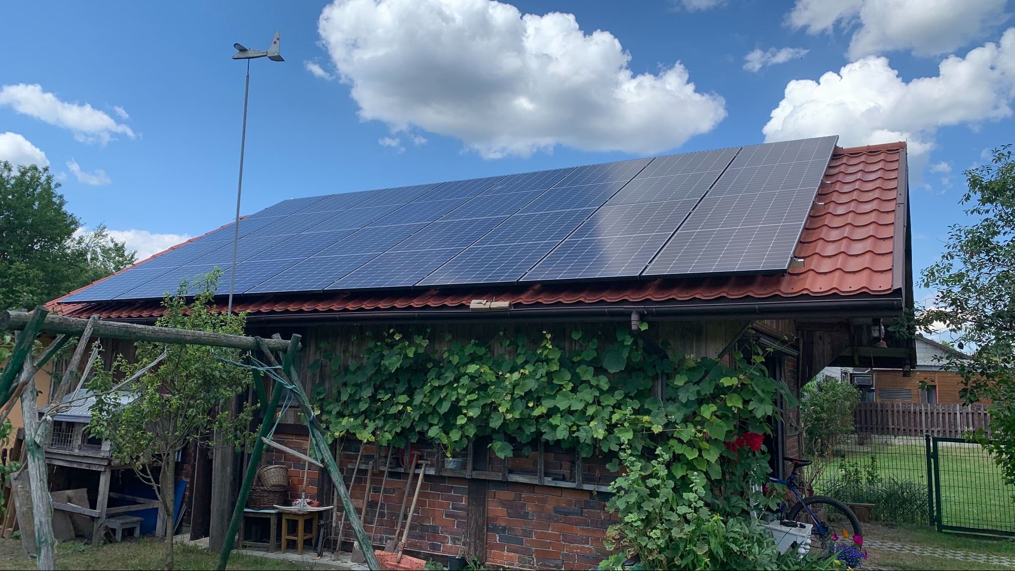 Panele słoneczne na dachu budynku gospodarczego z czerwoną dachówką, obrośniętego bluszczem, z wiatrowskazem w kształcie samolotu na szczycie.