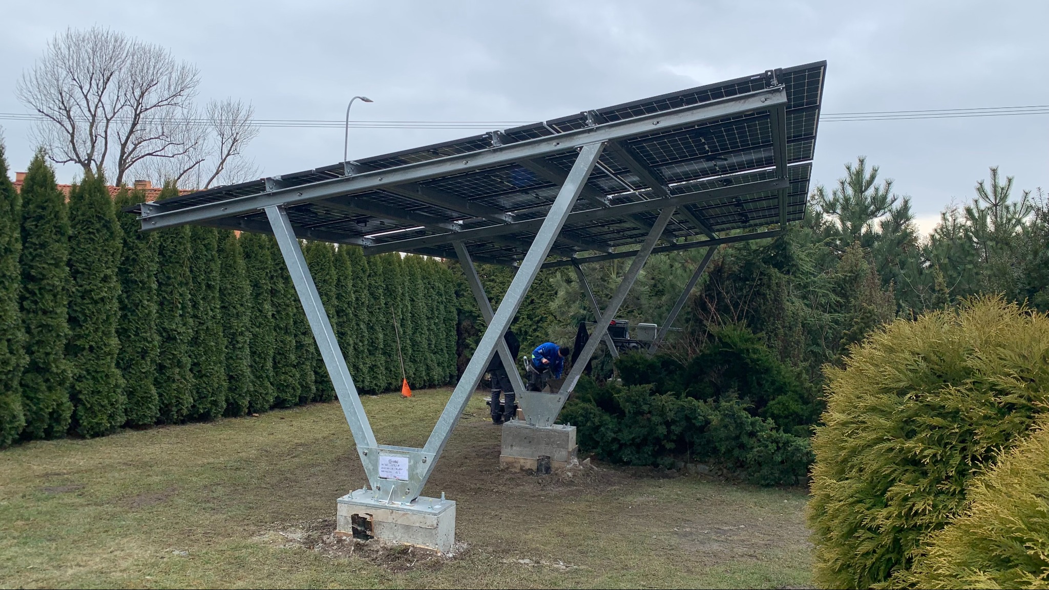 Montaż naziemnej instalacji fotowoltaicznej na metalowej konstrukcji w ogrodzie, widoczny pracownik w niebieskiej kurtce przy betonowych fundamentach.