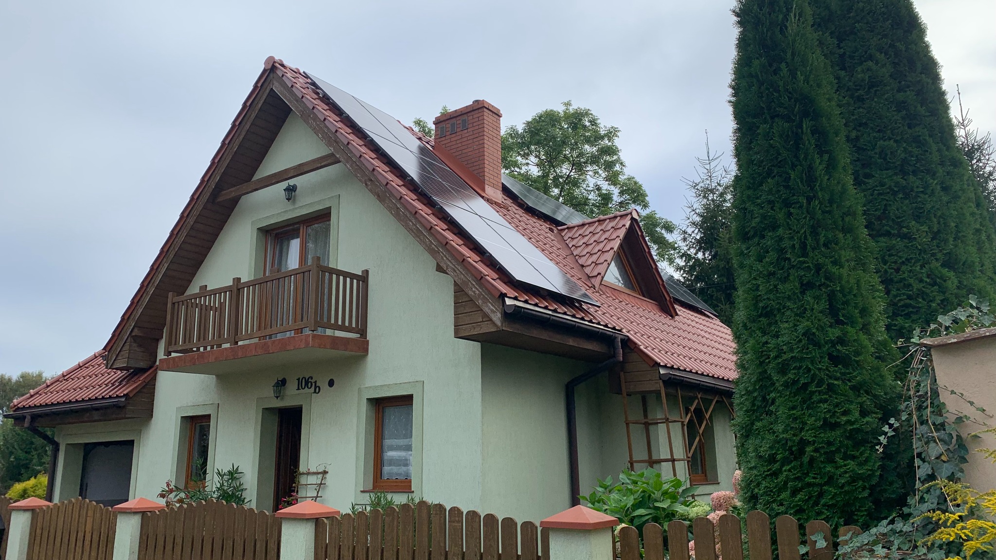 Dom jednorodzinny z zamontowanymi panelami słonecznymi na dachu pokrytym czerwoną dachówką, widoczne drewniane elementy wykończeniowe, balkon i zadbany ogród.