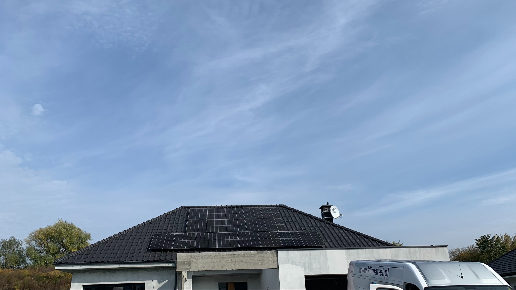 Panele słoneczne na dachu domu jednorodzinnego, widoczny samochód dostawczy firmy instalacyjnej z adresem strony internetowej, błękitne niebo z delikatnymi chmurami w tle.