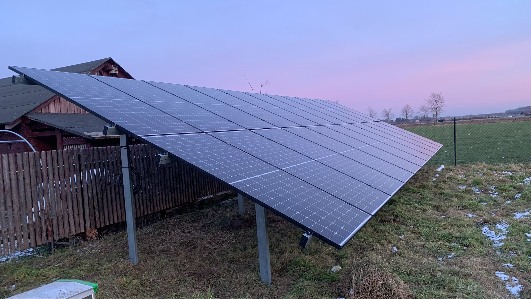 Panel fotowoltaiczny zamontowany na metalowej konstrukcji, widoczny na tle drewnianego budynku i łąki o zmierzchu.