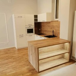 MEBLE-STYL DAWID ŁOPUSZYŃSKI - Jasna kuchnia z drewnianym blatem i wyspą z podświetlanymi półkami, widok na zabudowę kuchenną z piekarnikiem i czarnym zlewem, parkiet na podłodze.