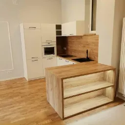 Jasna kuchnia z drewnianym blatem i wyspą z podświetlanymi półkami, widok na zabudowę kuchenną z piekarnikiem i czarnym zlewem, parkiet na podłodze.