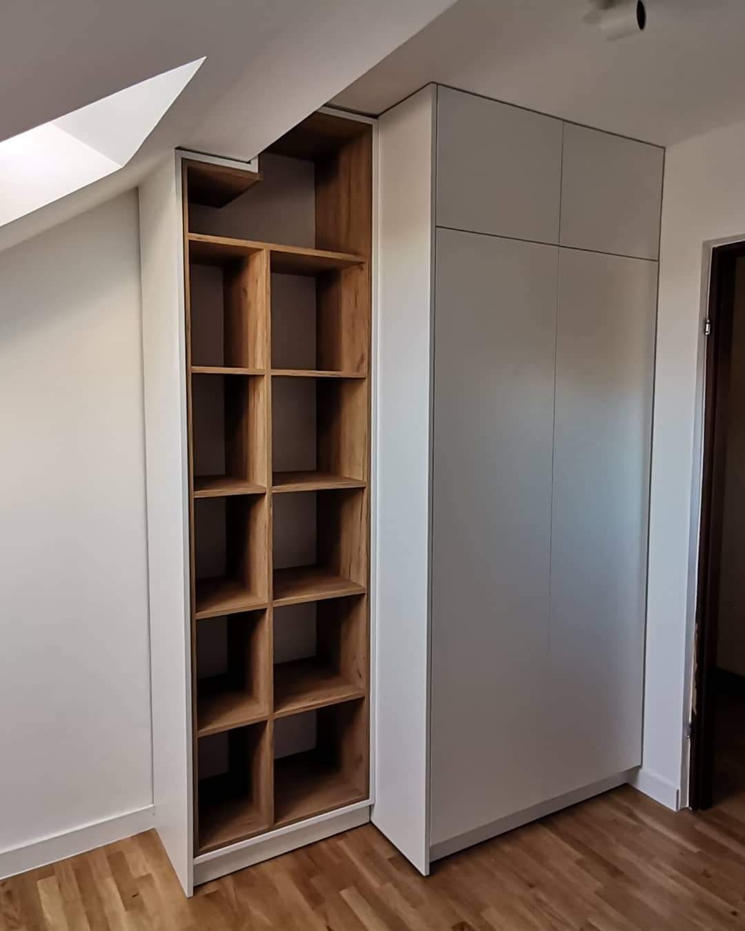 Szafa wnękowa z drewnianymi półkami w zabudowie pod skosem, fronty szafy w kolorze jasnoszarym, widoczna drewniana podłoga.