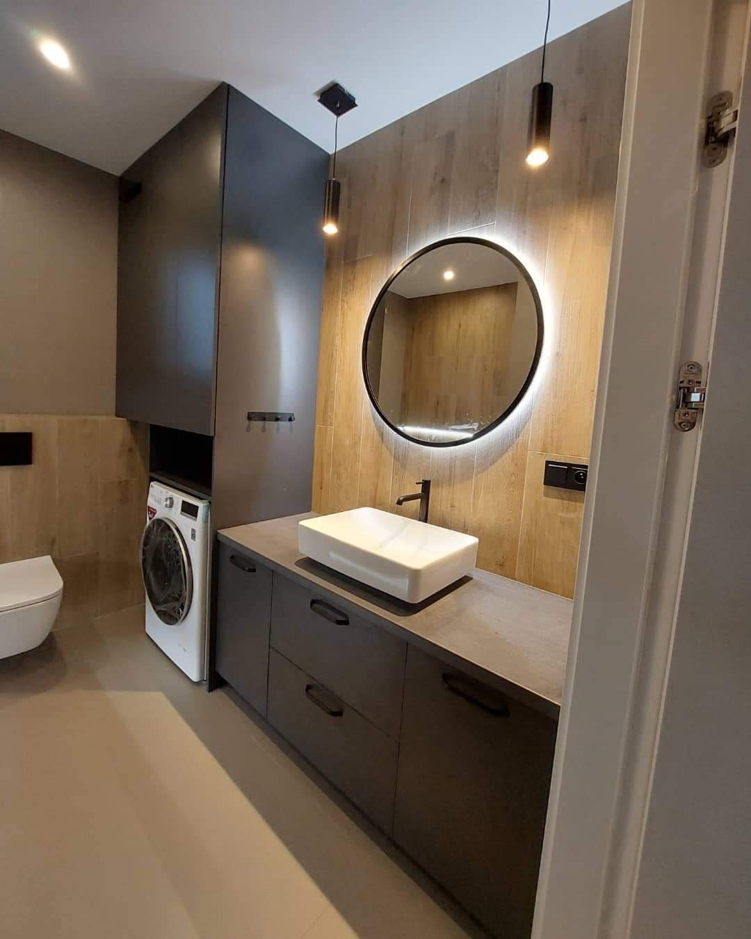 Nowoczesna łazienka z szarą zabudową meblową, umywalką nablatową, okrągłym lustrem podświetlanym LED i pralką w zabudowie.