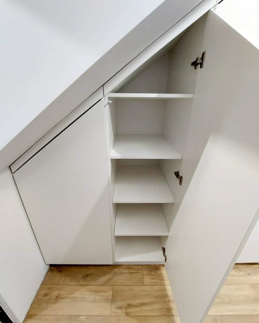 Biała szafa wnękowa z otwartymi drzwiami, dopasowana do skosu dachu, widoczne półki wewnętrzne i drewniana podłoga.