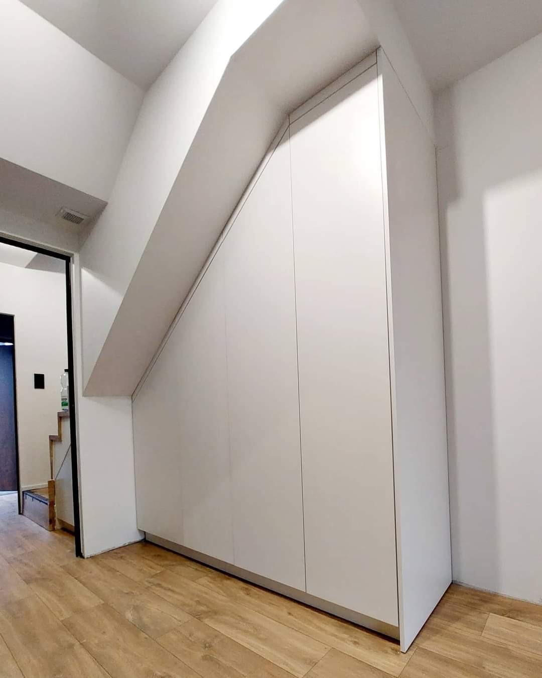 Biała zabudowa szafy wnękowej pod schodami, z panelami bez uchwytów, dopasowana do skosu, na drewnianej podłodze.