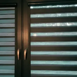 Okno z zamontowanymi roletami w kolorze turkusowo-szarym, z widocznymi poziomymi pasami zaciemniającymi i przepuszczającymi światło.