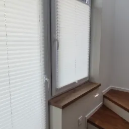 Białe plisy okienne zamontowane na dwóch oknach na klatce schodowej z drewnianymi stopniami, widoczne gniazdko elektryczne pod parapetem.