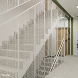 Klatka schodowa w Gdyni z białymi schodami i ażurową balustradą z siatki, widok z dołu na górę, z zielonym akcentem kolorystycznym na ścianie i rośliną w donicy.