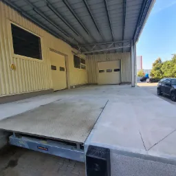 Rampa załadunkowa z platformą podnoszoną, widoczne dwie bramy segmentowe i fragment samochodu osobowego na zewnątrz budynku.