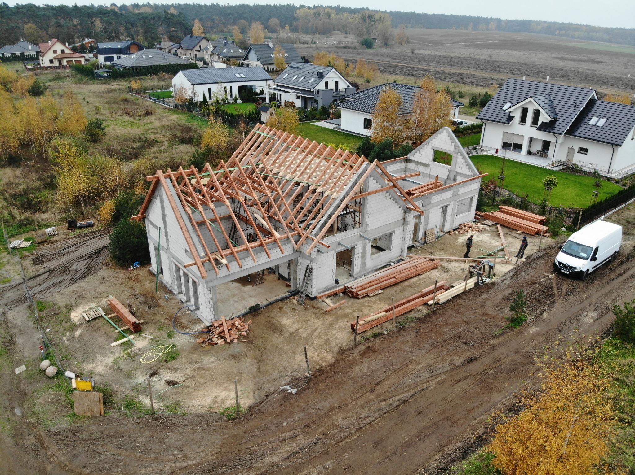 Budowa domu jednorodzinnego z pustaków, widok z góry na konstrukcję dachu w trakcie montażu, obok leżą przygotowane elementy drewniane, w tle zabudowa osiedlowa i las.