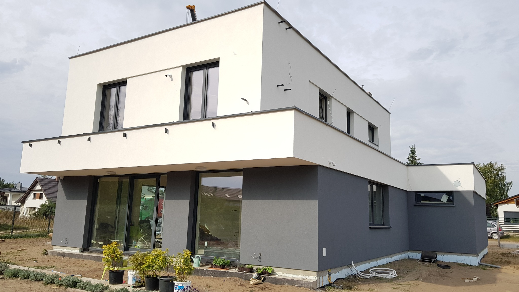 Nowoczesny dwukondygnacyjny dom z płaskim dachem, z szarą elewacją na parterze i białą na piętrze, duże okna, widoczne rośliny w donicach przed wejściem, teren wokół w trakcie zagospodarowywania.