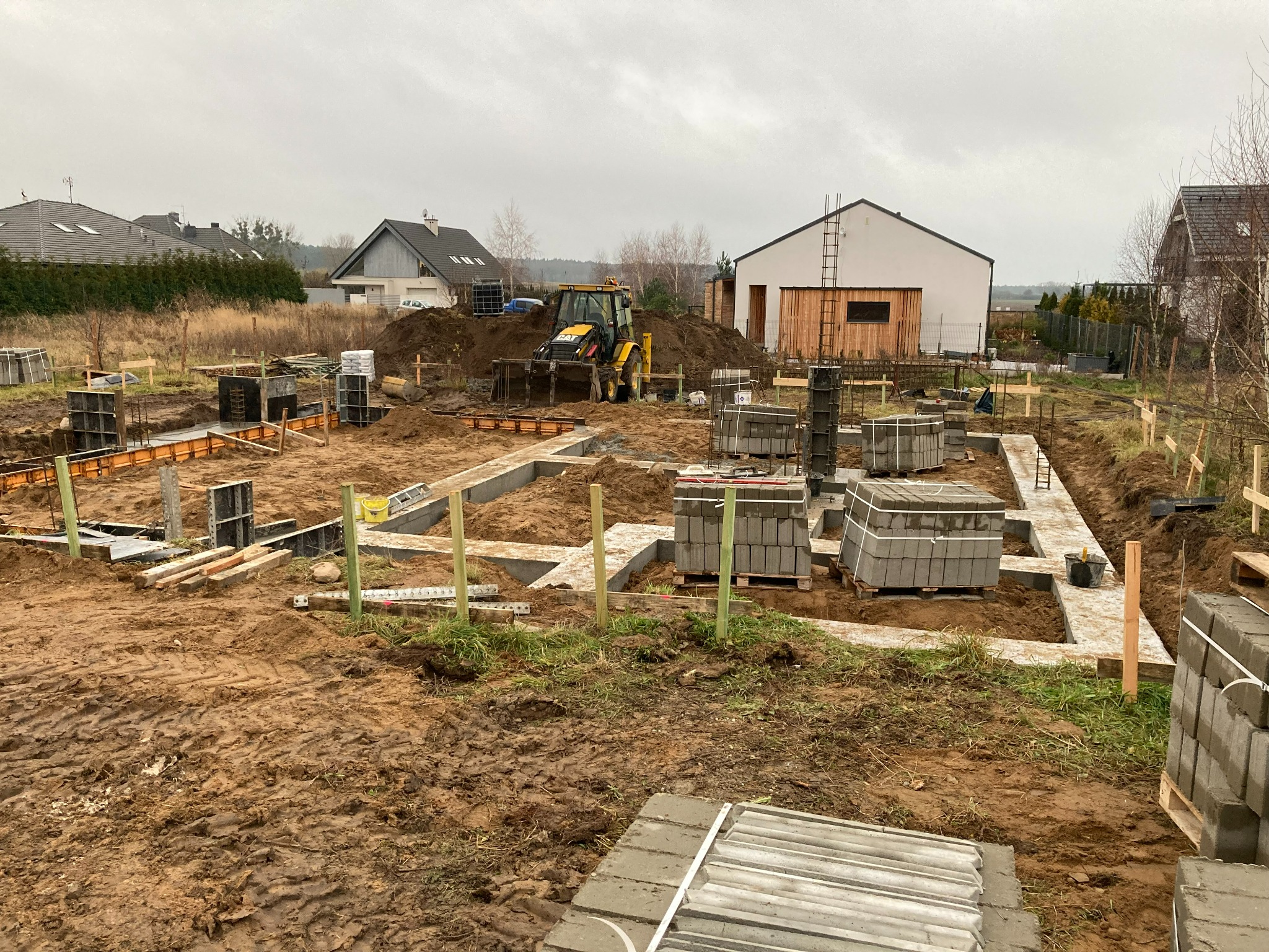 Fundamenty domu w trakcie budowy, widoczne szalunki, betonowe bloczki i koparka w tle, pochmurny dzień.