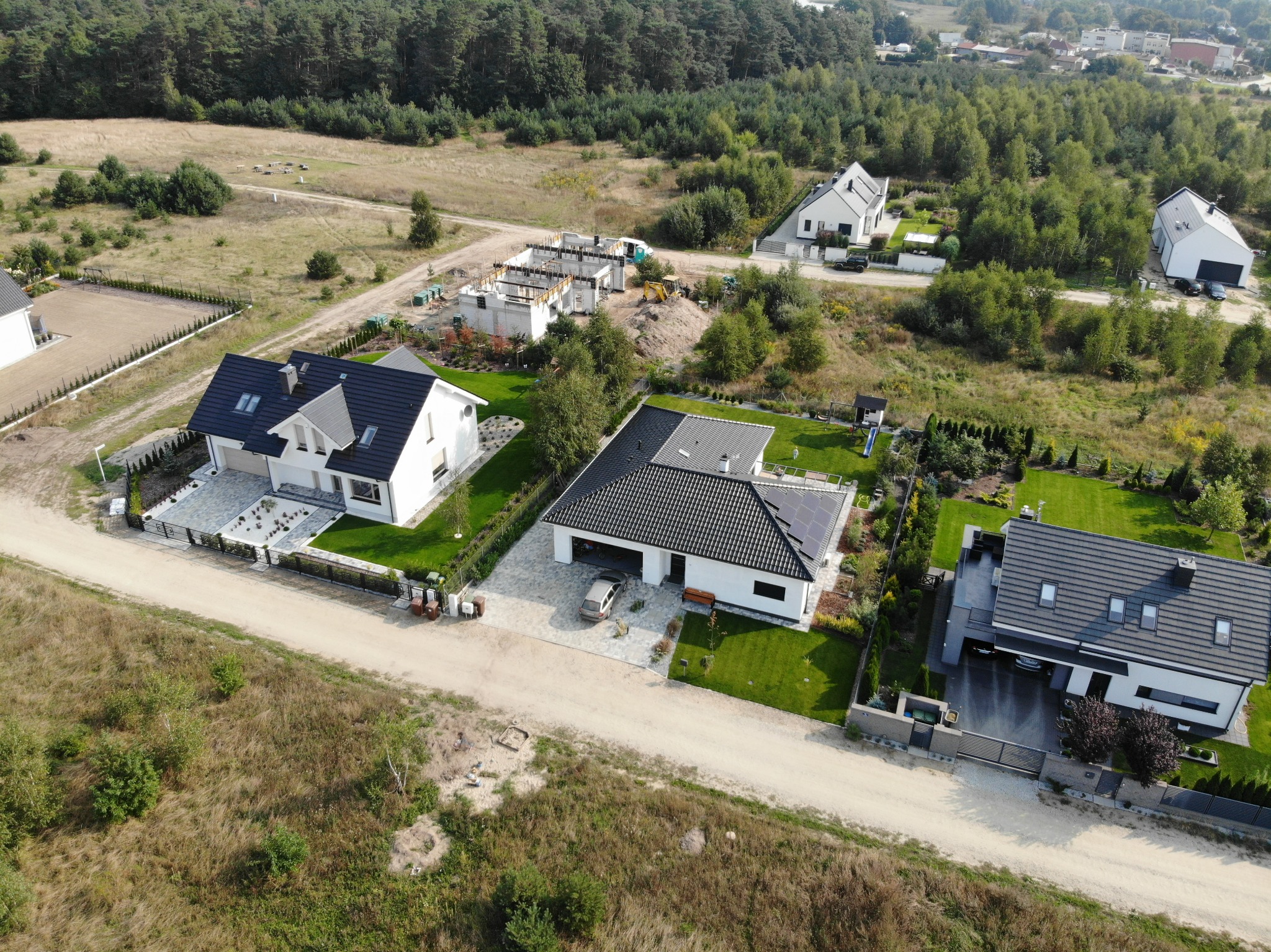 Widok z lotu ptaka na osiedle domów jednorodzinnych w różnym stadium zaawansowania budowy, od fundamentów po ukończone budynki z zagospodarowanym terenem.