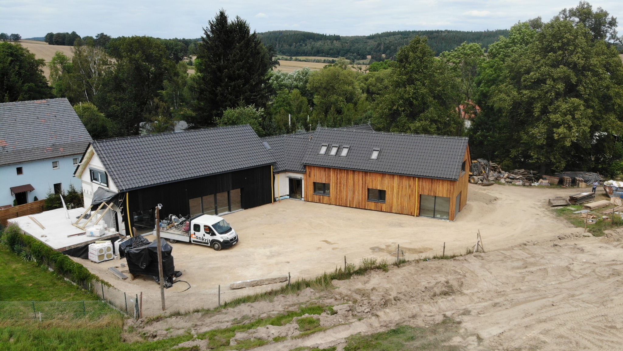 Nowoczesny dom z drewnianą elewacją i czarnym dachem, częściowo wykończony, z widocznym samochodem dostawczym i materiałami budowlanymi na posesji, w tle las