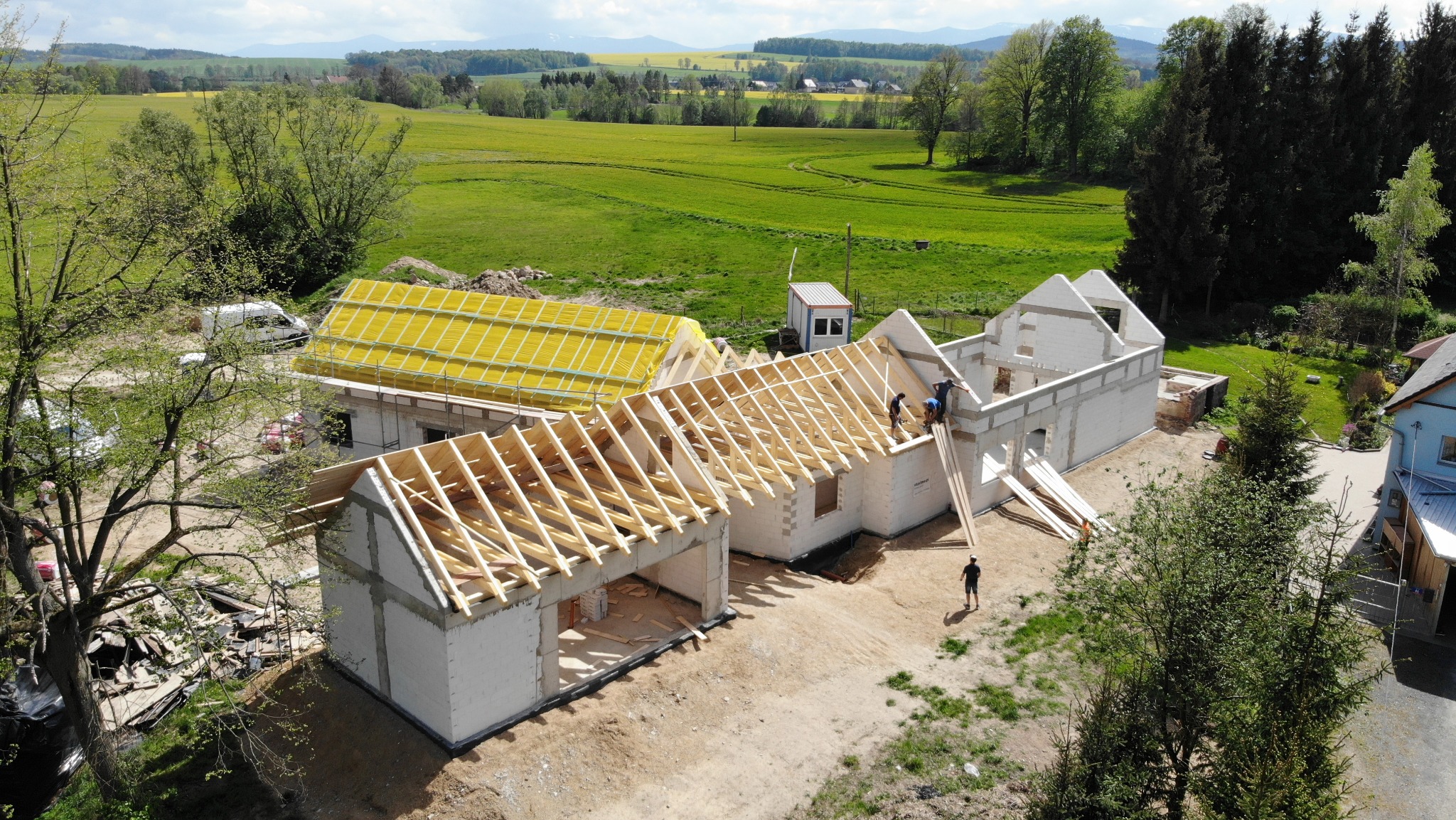Budowa domu z białych bloczków, widok z góry na konstrukcję dachu z drewnianych belek, dwóch pracowników na dachu, w tle zielone pola i lasy.