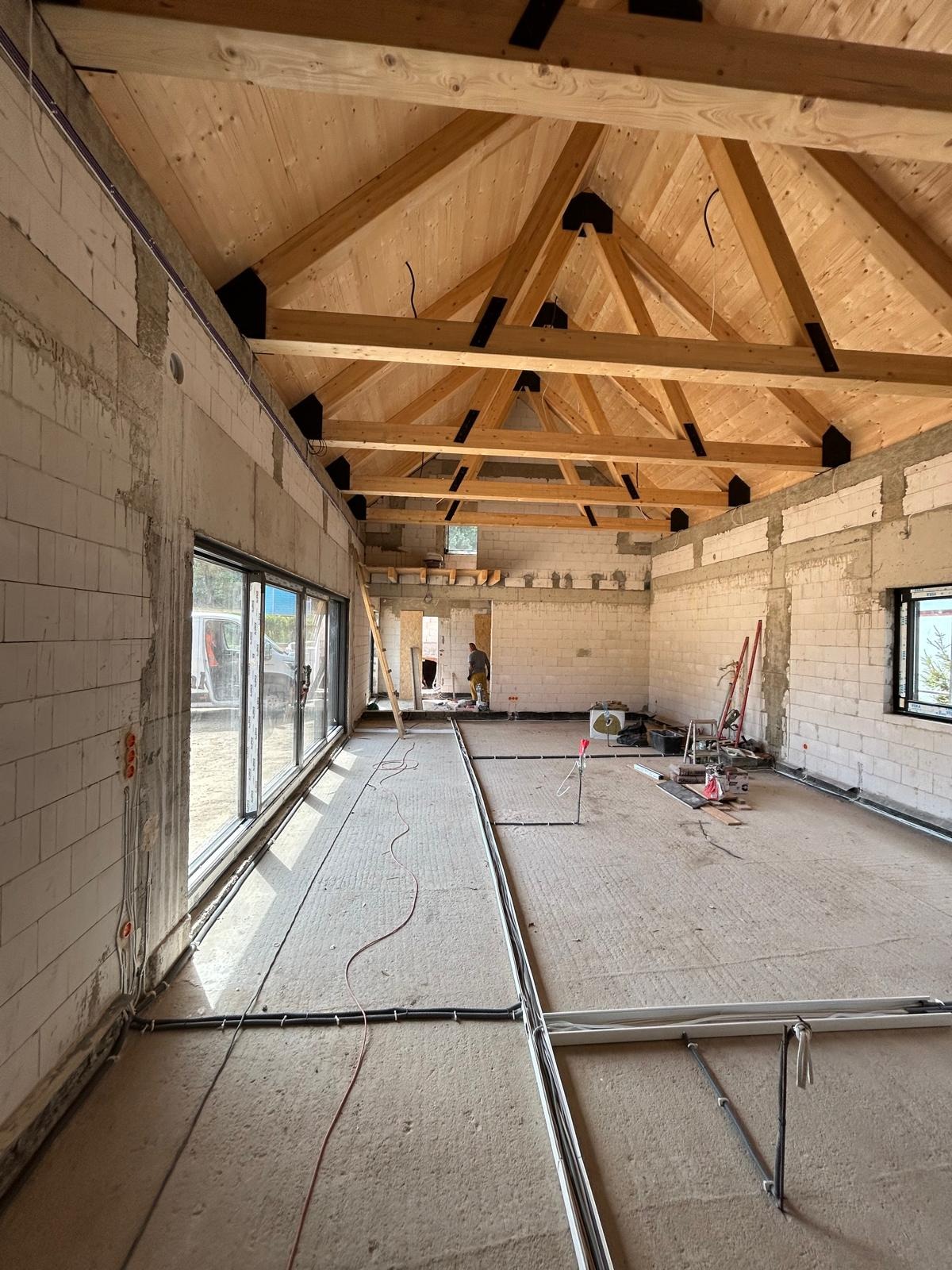 Wnętrze domu w stanie surowym otwartym z widoczną konstrukcją drewnianą dachu, ścianami z białych bloczków oraz instalacją elektryczną pod podłogą.