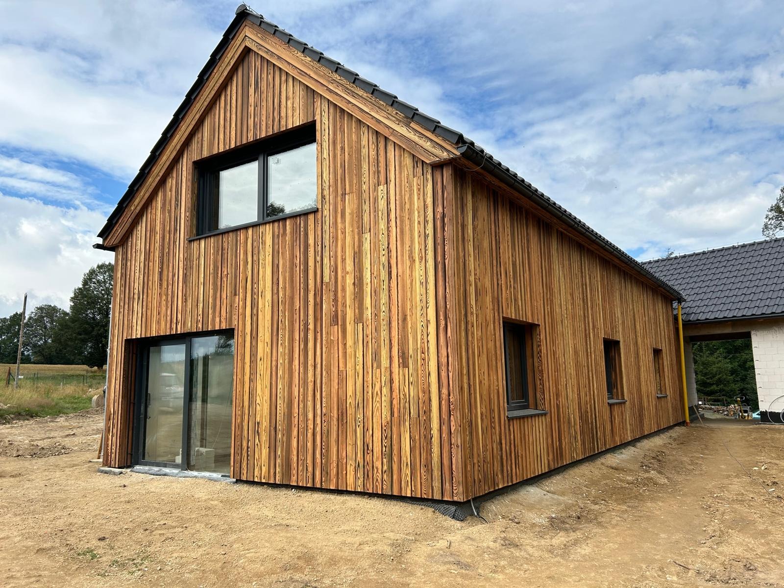 Nowoczesny dom z elewacją z pionowych desek o ciepłym, miodowym odcieniu, czarne okna i dachówka, widok z boku na tle błękitnego nieba.