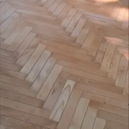 Jasna podłoga z parkietu ułożonego we wzór jodełki, oświetlona naturalnym światłem słonecznym.