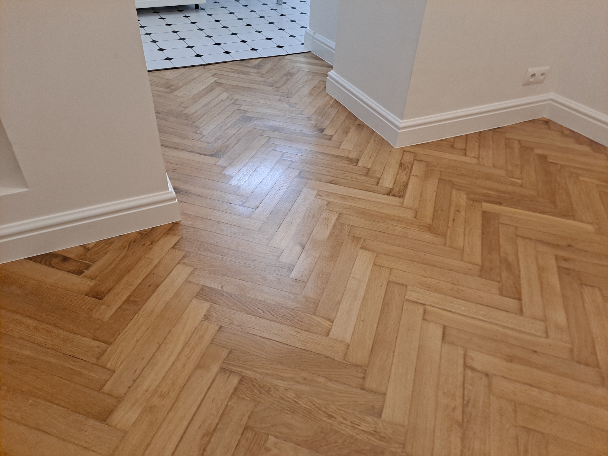 Jasna drewniana podłoga ułożona w jodełkę, widoczna z perspektywy wejścia do pomieszczenia z białą podłogą w czarno-białą szachownicę.