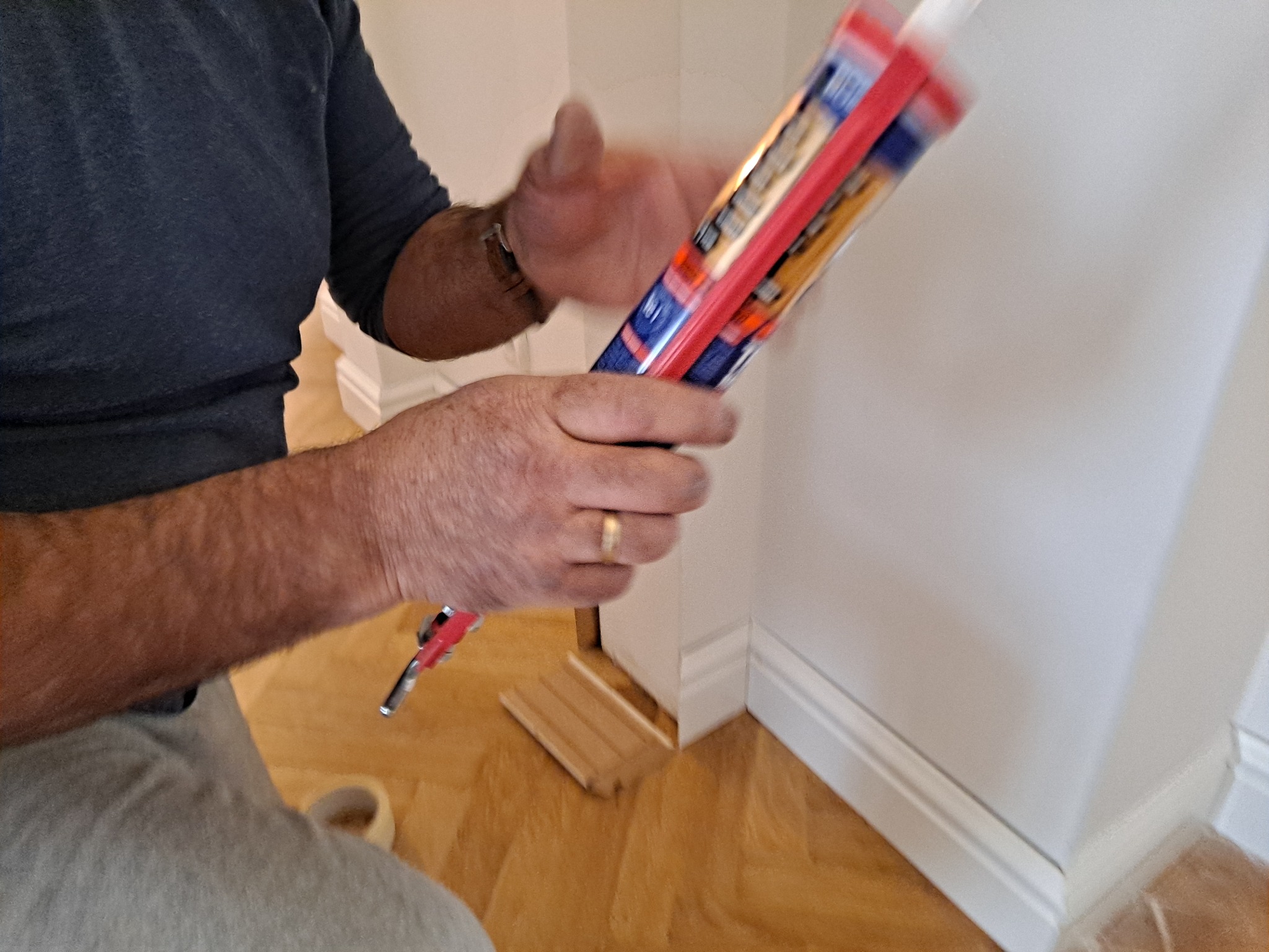 Montaż listew przypodłogowych na parkiecie, widoczny pistolet do kleju i fragment dłoni majstra z obrączką.